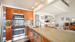 A modern kitchen with granite countertops, stainless steel appliances, wooden cabinets, and a large island with a sink. - Pali Ke Kua #222 - Breathtaking Ocean Views! (Princeville)