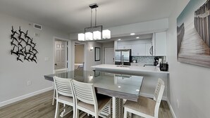 Uma cozinha moderna com armários brancos, uma ilha central com bancada de vidro e uma área de refeições com cadeiras brancas.