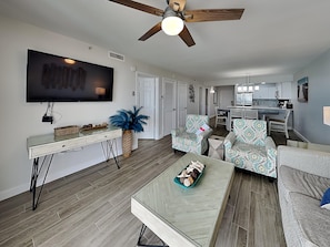 A modern living room with a flat-screen TV, a ceiling fan, and a dining area with a kitchen in the background. - Pelican Beach #2017 | Beachfront, Views, Amenities (Destin)