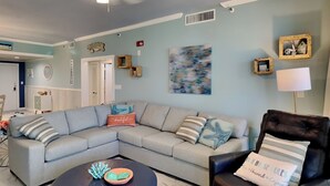 A living room with a sectional sofa, a round ottoman, a black leather armchair, and a wall-mounted lamp. - A Joyful Breeze | Beachfront, Pool, Grills, Fun! (Navarre Beach)