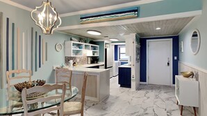 A modern kitchen with white cabinets, a marble floor, and a dining area with a glass table and wooden chairs. - A Joyful Breeze | Beachfront, Pool, Grills, Fun! (Navarre Beach)