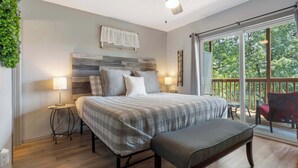 Quarto com cama, mesas de cabeceira, um banco, ventilador de teto e varanda com vista para árvores.
