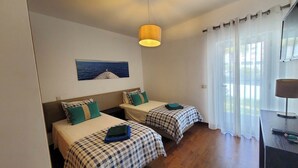 A room with two beds, a ceiling lamp, a painting of a boat on water, and a window with curtains. - House (Albufeira)