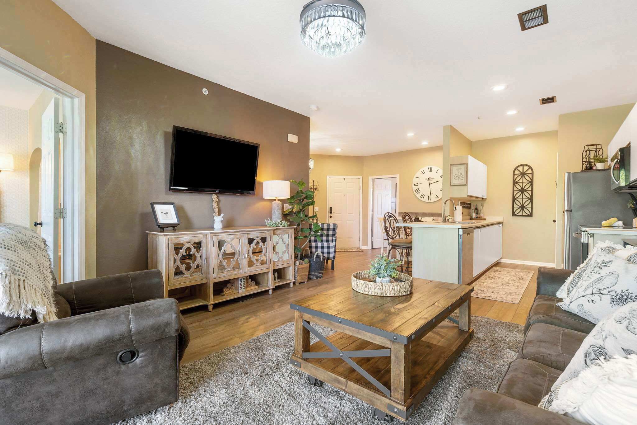 Una sala de estar con un sofá, una mesa de centro y un televisor. Hay una zona de cocina con armarios y un refrigerador.