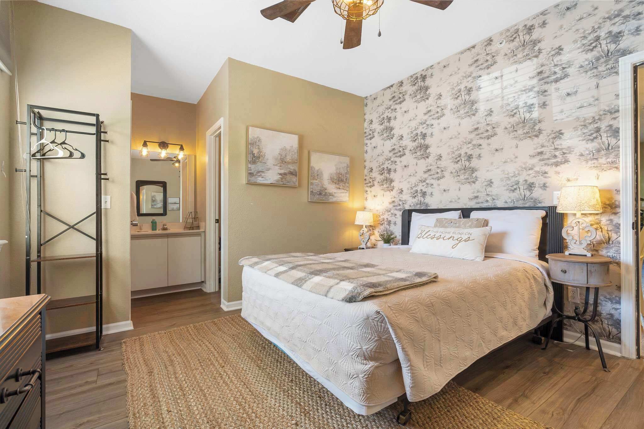 A bedroom with a bed, a nightstand, a lamp, a dresser, a mirror, and a hanging rack.