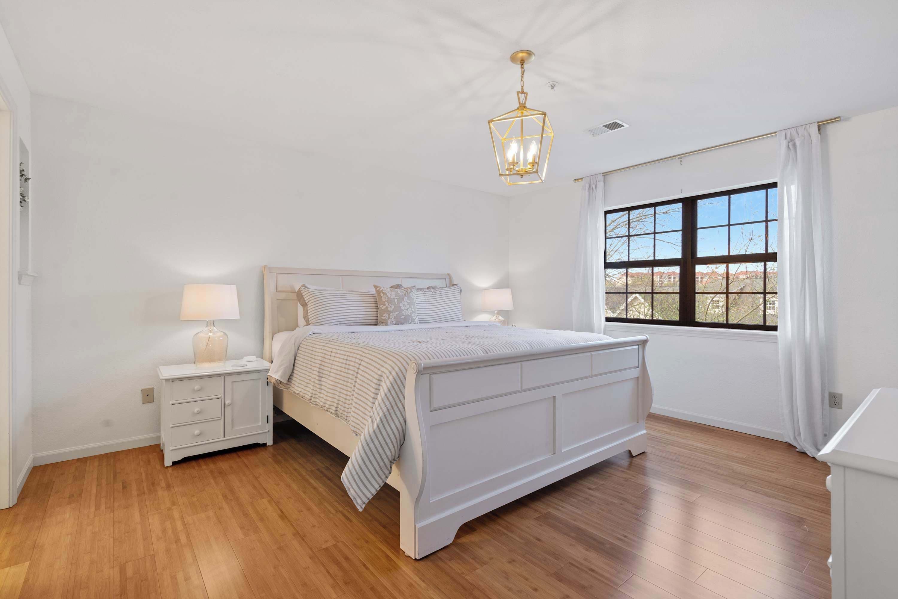 Un dormitorio con una cama grande, una mesita de noche con lámpara, una ventana con cortinas y un candelabro.