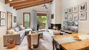 A spacious living room with a fireplace, a large window, and a dining area with a wooden table. - Gold Bend Bliss (Truckee)