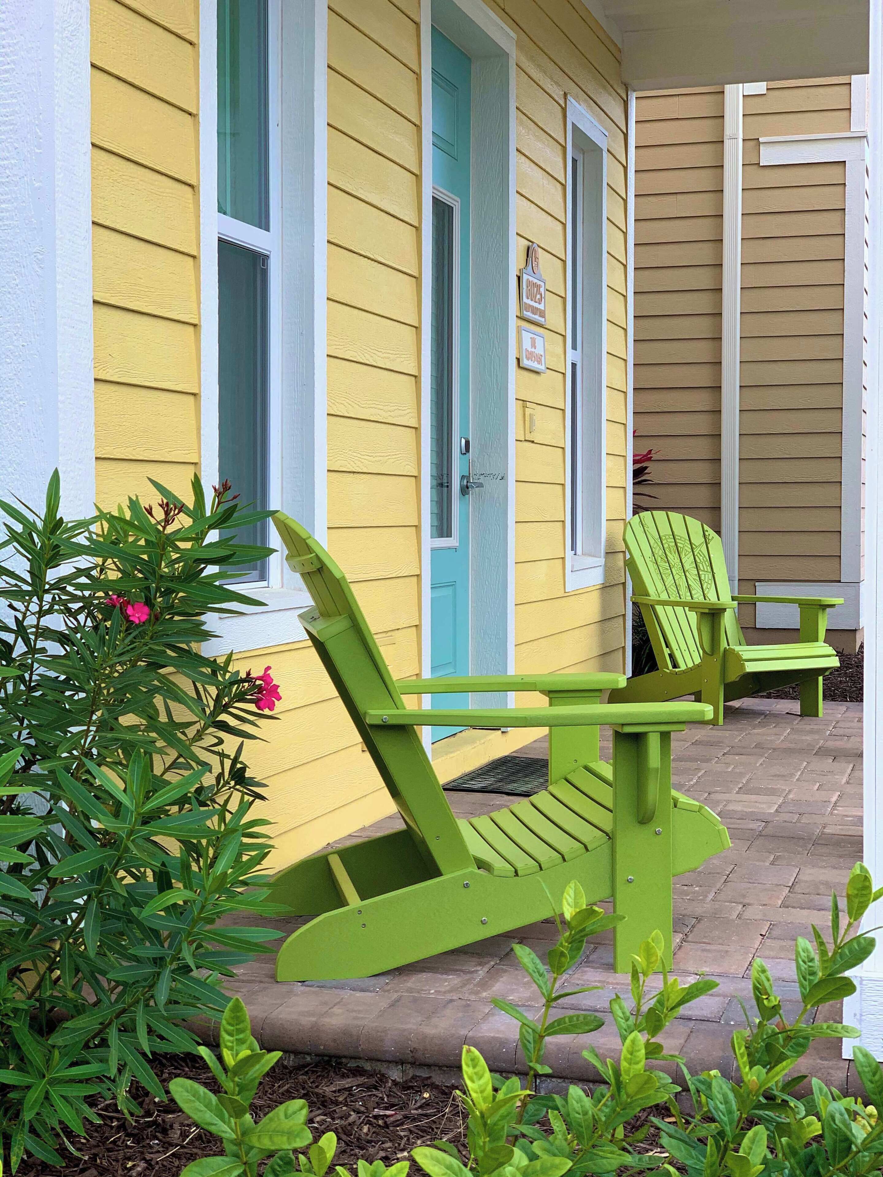 A yellow building with a turquoise door and windows, green Adirondack chairs, and a small garden with pink flowers.