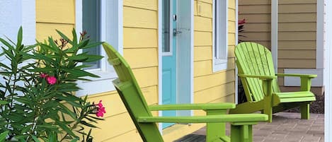 A yellow building with a turquoise door and windows, green Adirondack chairs, and a small garden with pink flowers.