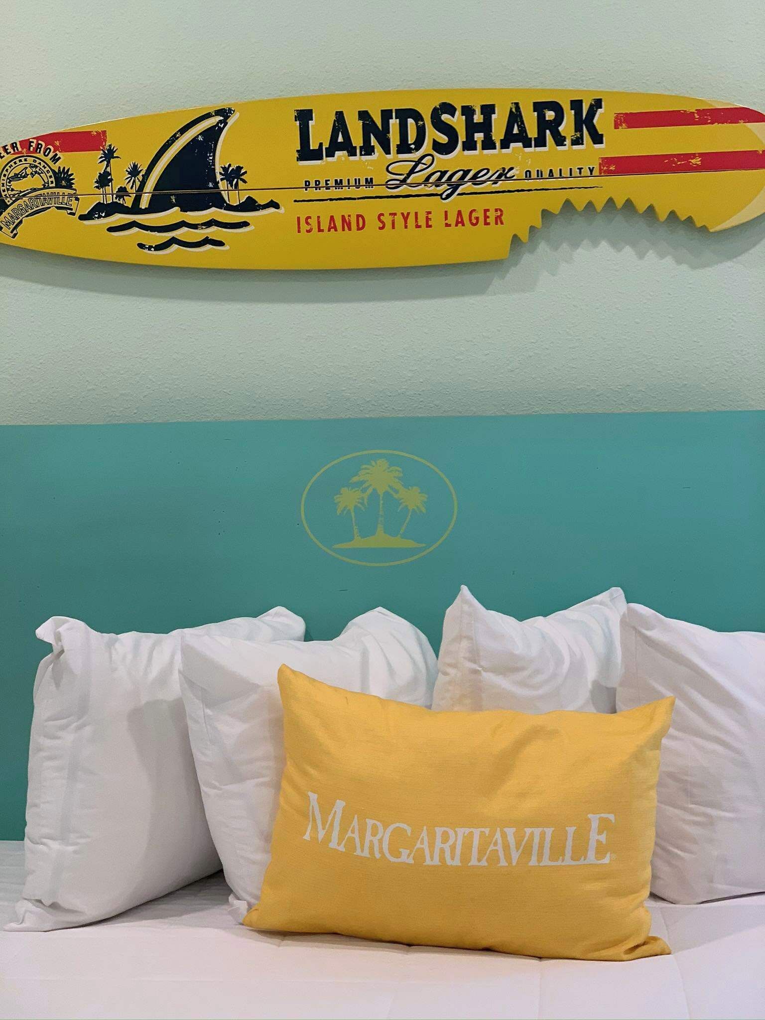 A bed with white and yellow pillows, a sign above the bed, and a teal wall with a logo.