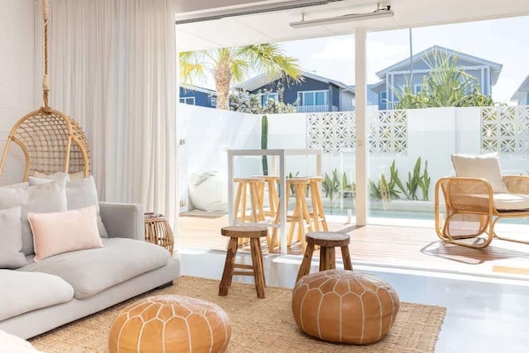 A modern living room with a sofa, ottomans, and a hanging chair. There is a balcony with a table and chairs, and a view of houses and greenery.