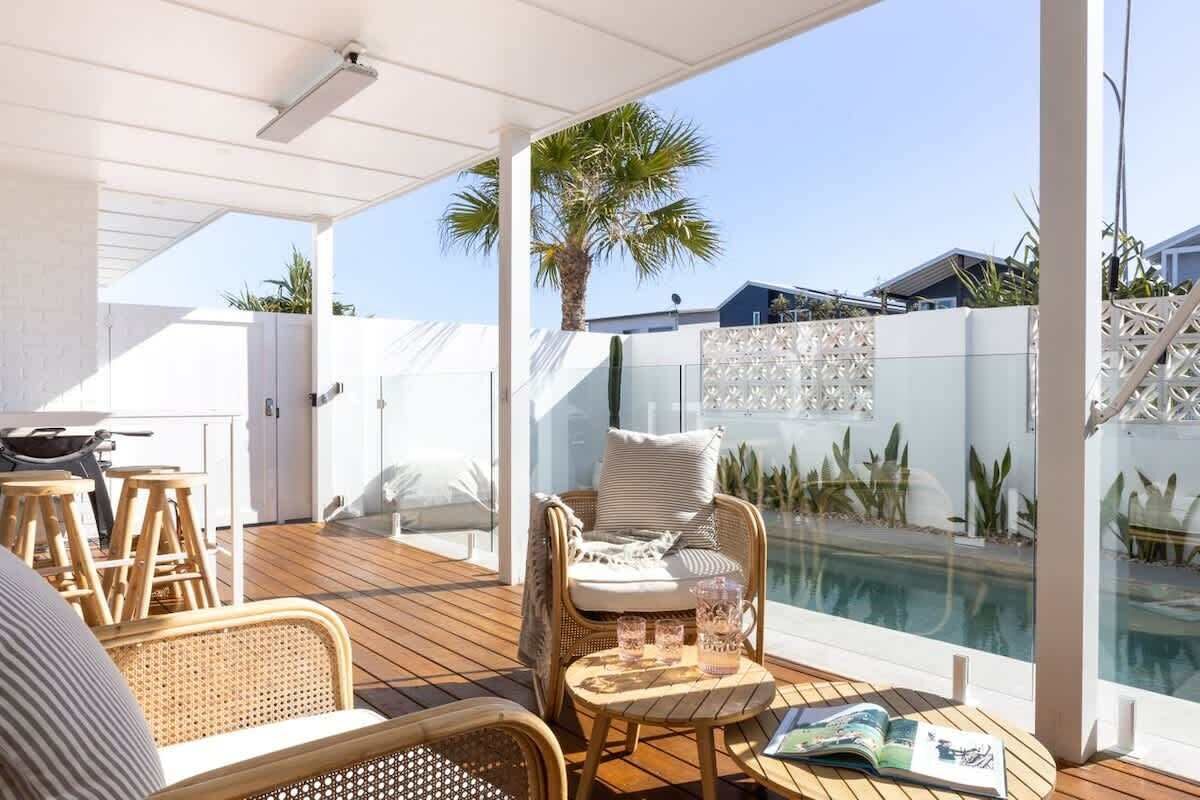 An outdoor deck with wicker furniture, a glass table, and a pool in the background.
