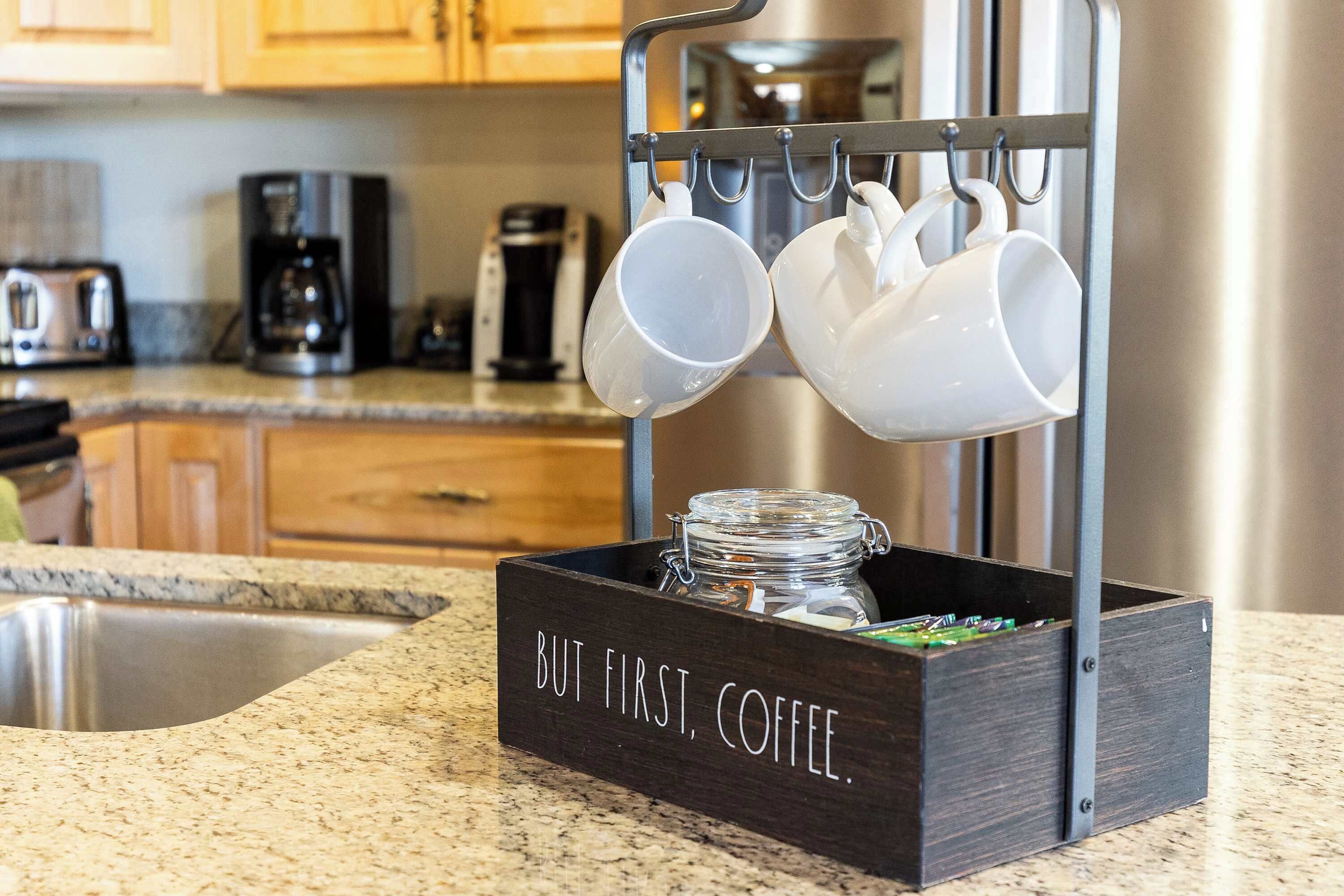 A kitchen counter with a coffee station featuring a wooden box labeled "BUT FIRST, COFFEE."