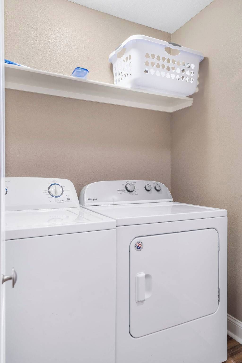 Une buanderie avec deux machines à laver blanches, un panier blanc posé sur une étagère et un mur de couleur beige.