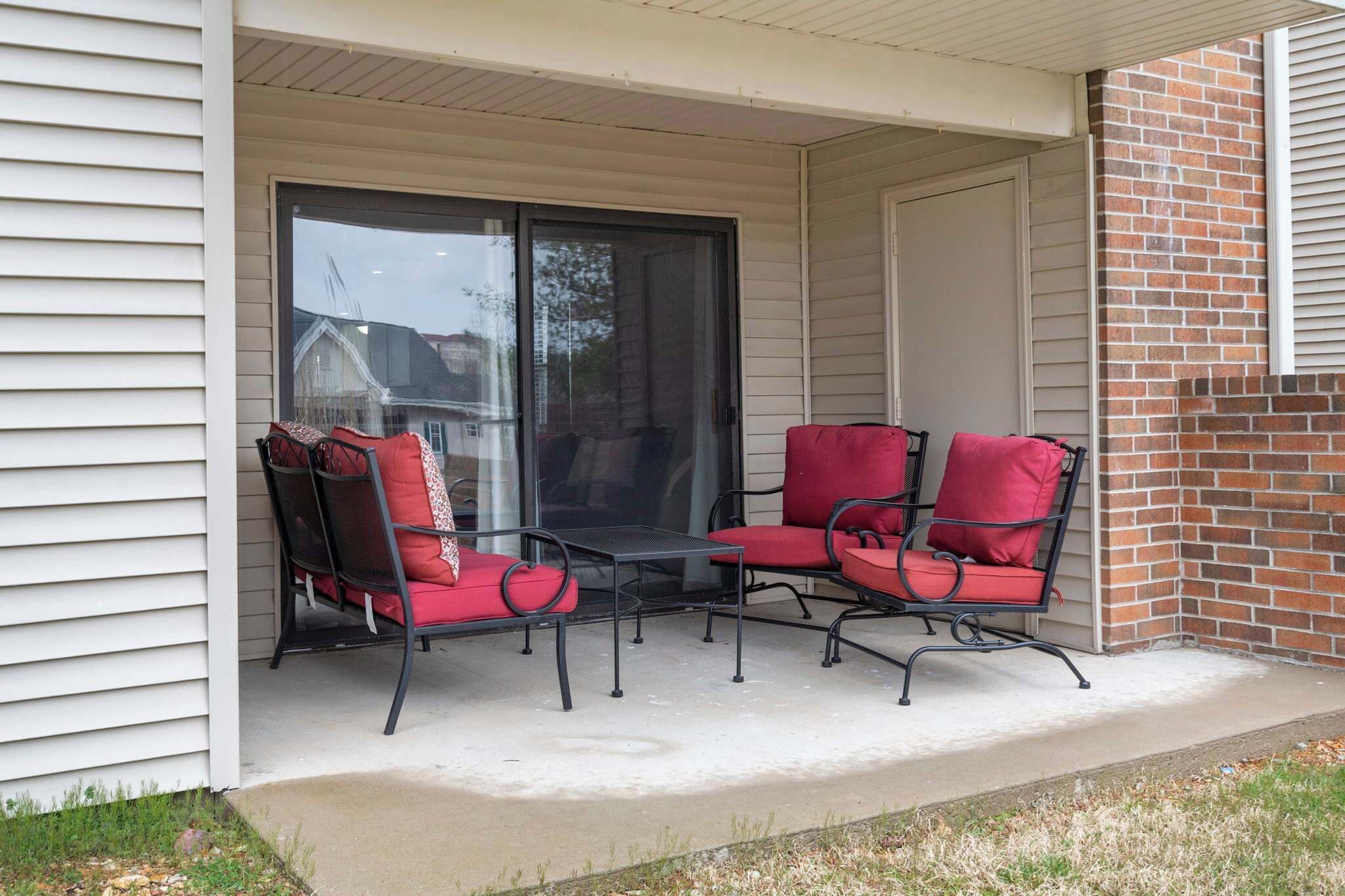 A covered patio with two red cushioned chairs and a small table, adjacent to a sliding glass door leading to a brick wall.