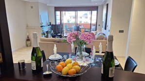 A dining area with a table set for two, featuring a bottle of wine, a vase with flowers, and a fruit basket.
