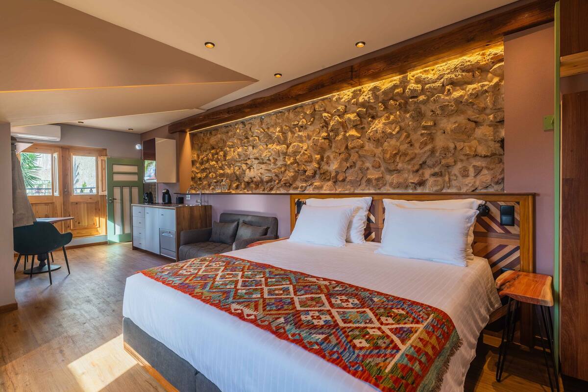 Una habitación de hotel moderna con una cama grande, una pared decorativa de piedra, un pequeño rincón de estar y una kitchenette.
