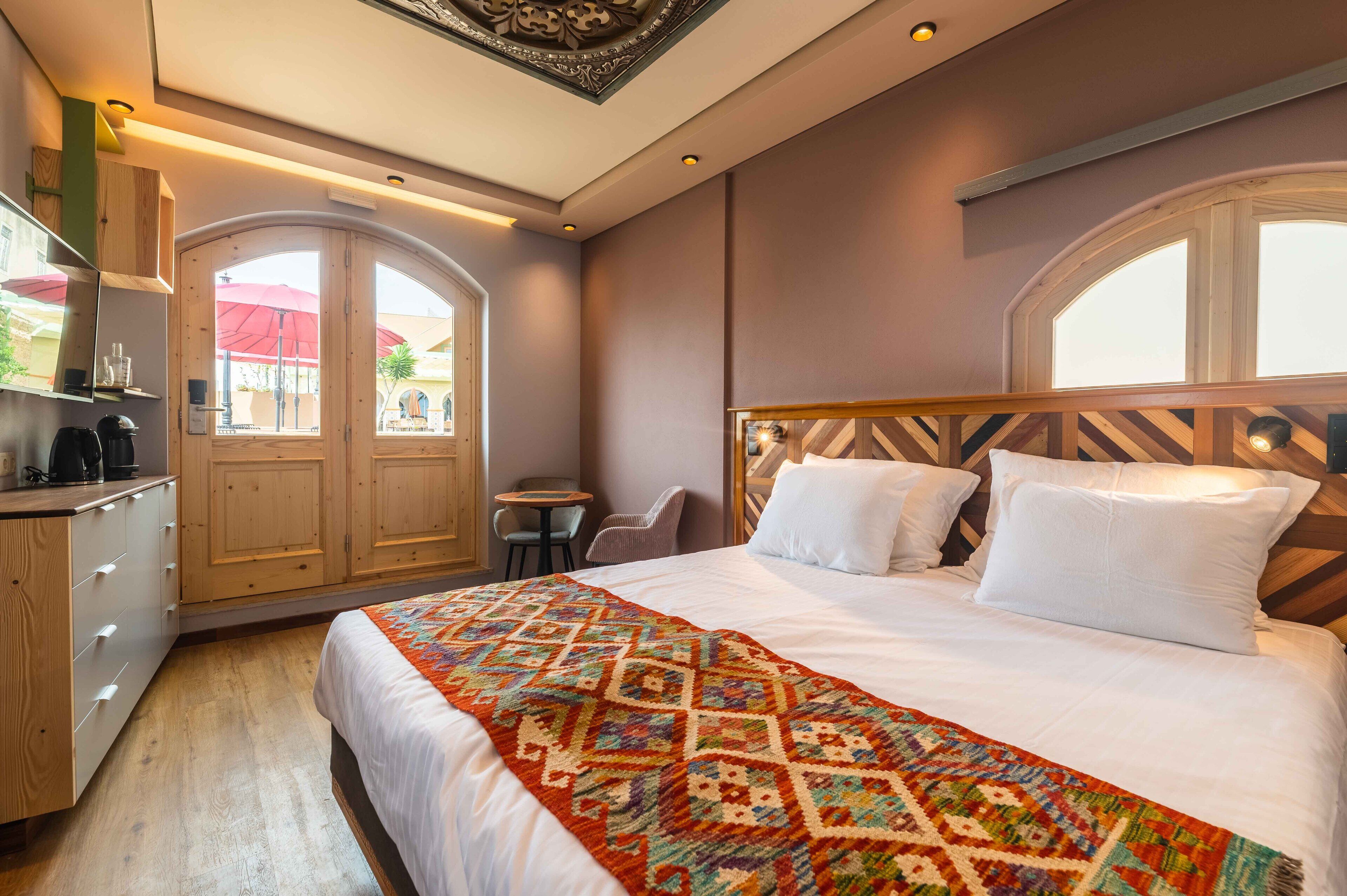 Une chambre d’hôtel avec un grand lit, une tête de lit en bois, une petite table et une chaise. Il y a une fenêtre donnant sur l’extérieur.