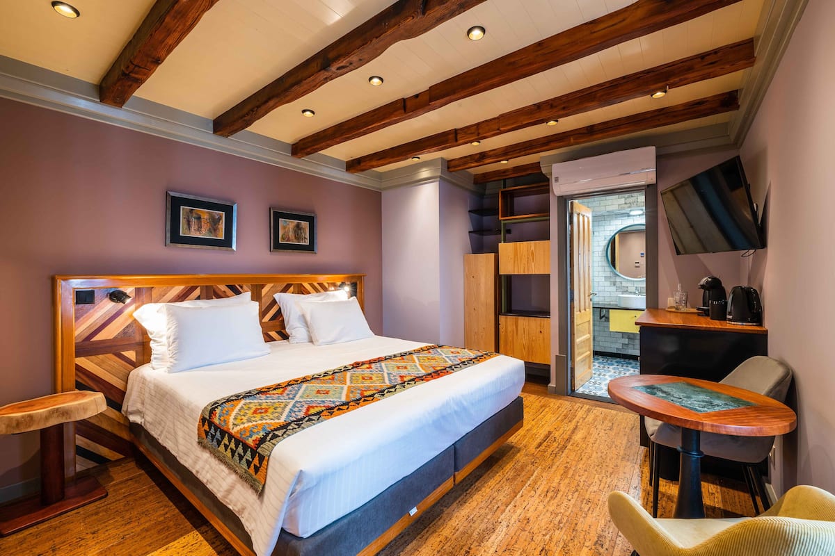 Habitación de hotel con una cama de madera, una mesita pequeña, una silla y vistas a la cocina y al baño a través de una puerta abierta.