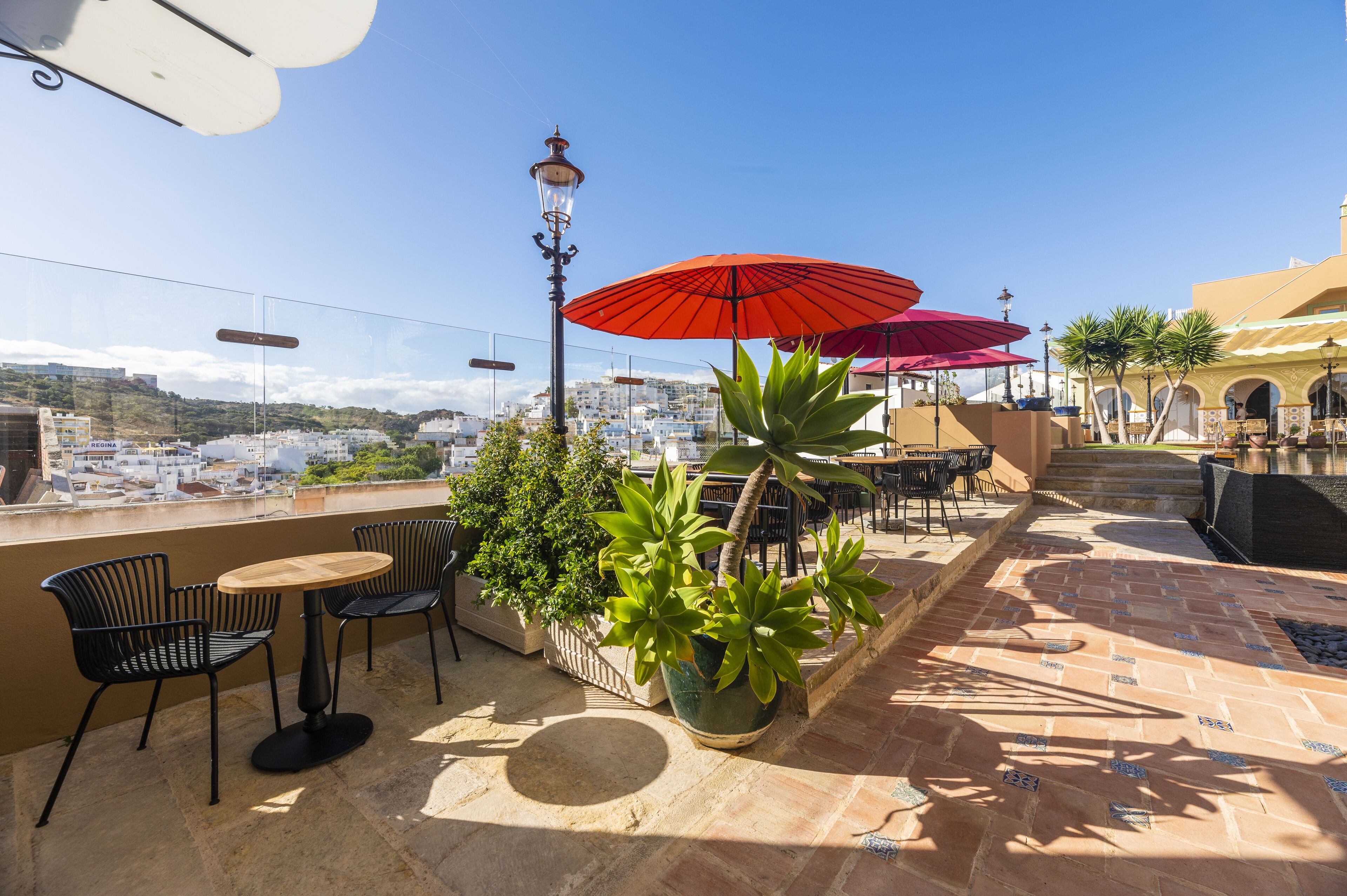 An outdoor patio with tables and chairs, red umbrellas, and potted plants.