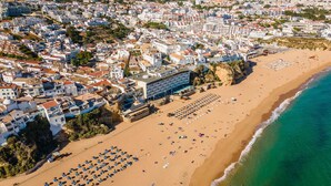 An aerial view of a coastal town with a wide sandy beach, numerous beach umbrellas, and a hotel with a large building adjacent to the beach. - Vila Origens Boutique Hotel Albufeira (Albufeira)