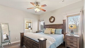Une chambre avec un lit en bois, un ventilateur de plafond, une table de chevet avec une lampe, un miroir et une fenêtre avec des rideaux.