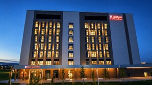Un hôtel moderne, avec plusieurs étages, des fenêtres illuminées et une enseigne Hilton Garden Inn bien visible.