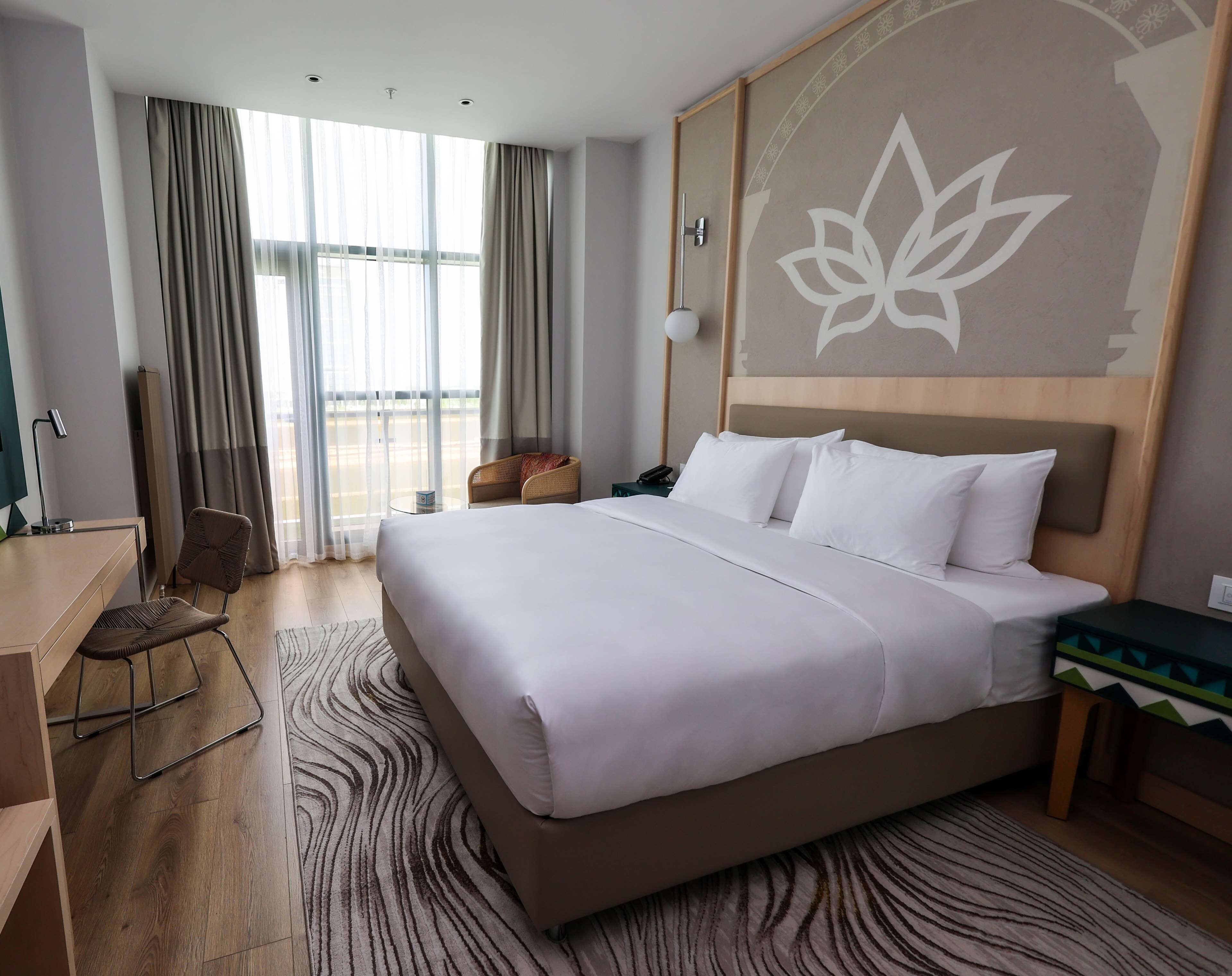 A hotel room with a large bed, a desk, a chair, a view of the outdoors, and a decorative wall piece.