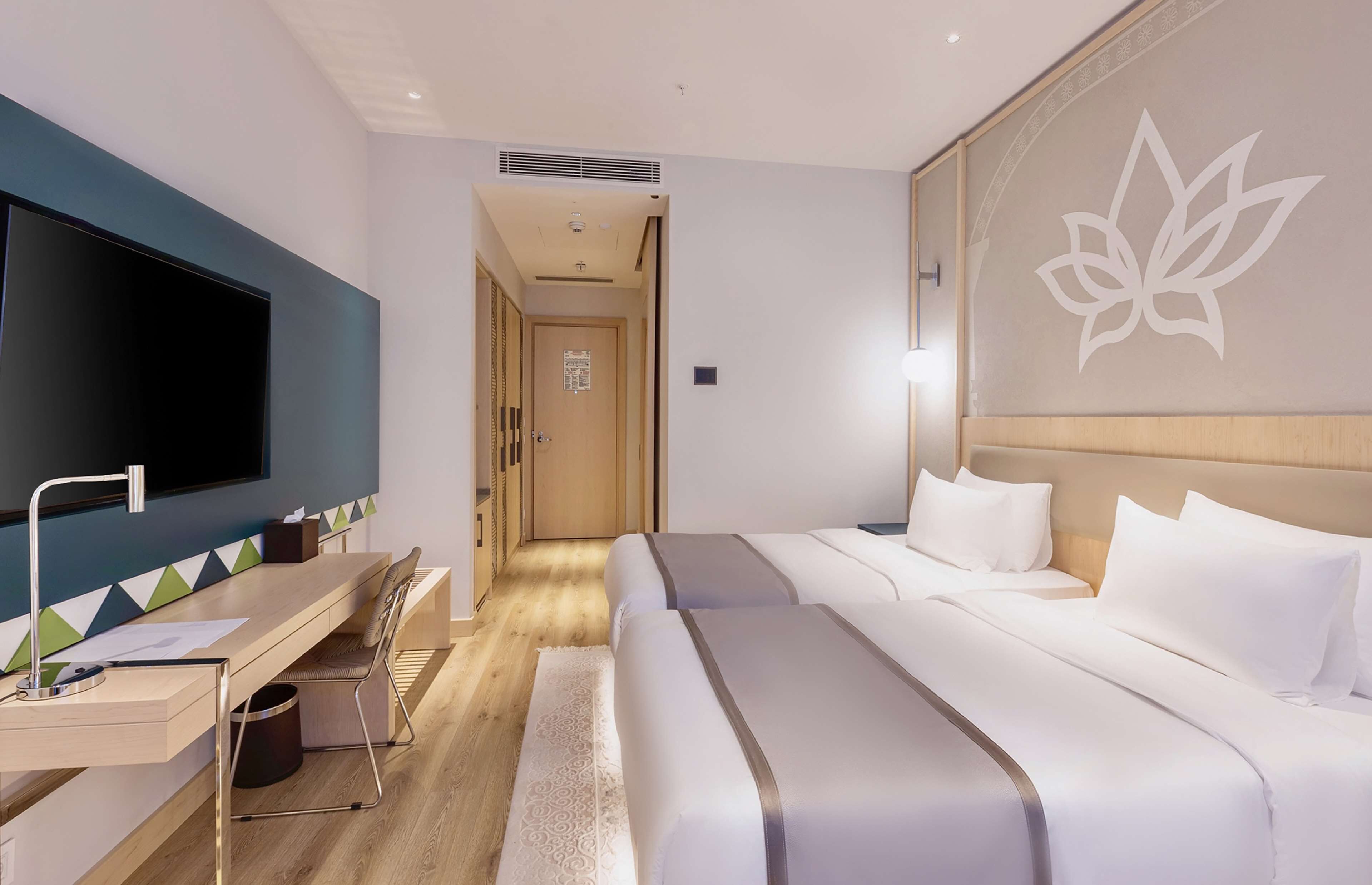 A hotel room with a bed, a desk with a chair, a television, and a wall with a lotus flower design.