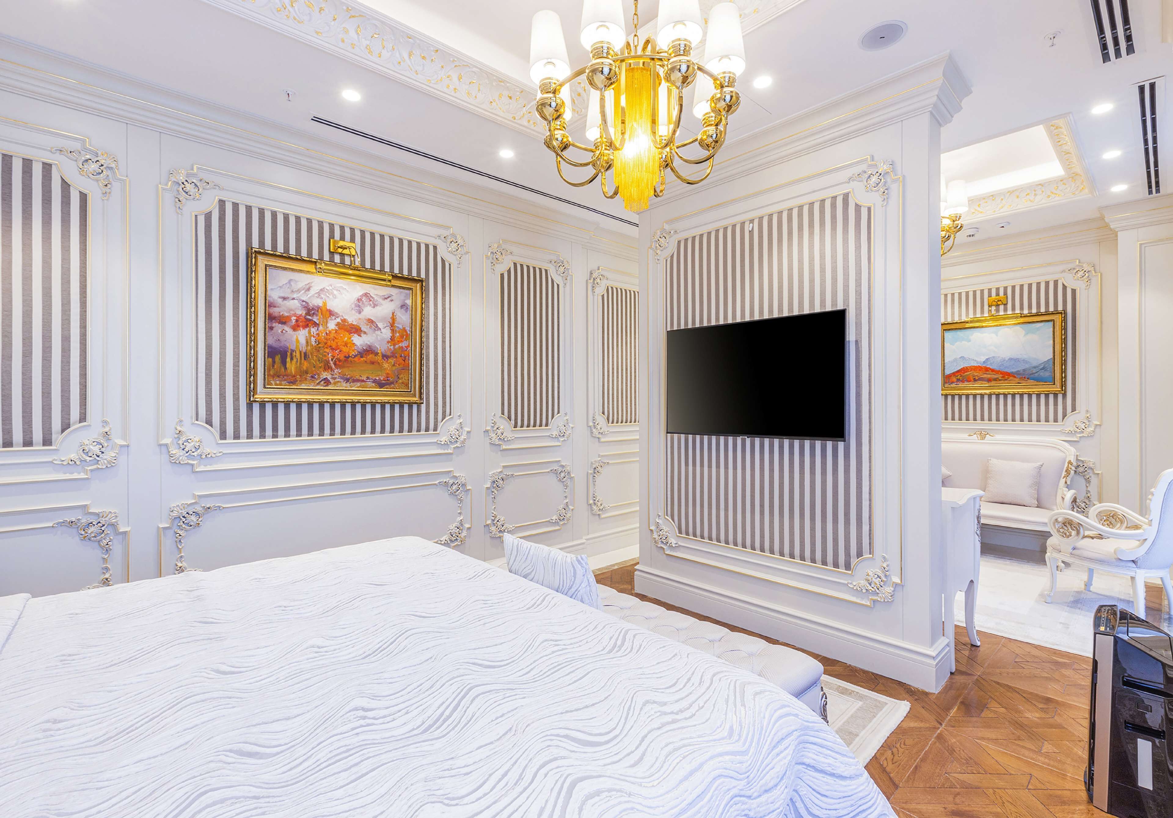 A luxurious bedroom with a large bed, a flat-screen TV, ornate wall decorations, and a chandelier.