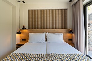 A modern hotel room with a large bed, wooden headboard, two bedside tables with lamps, and a view of the outdoors through a large window.
