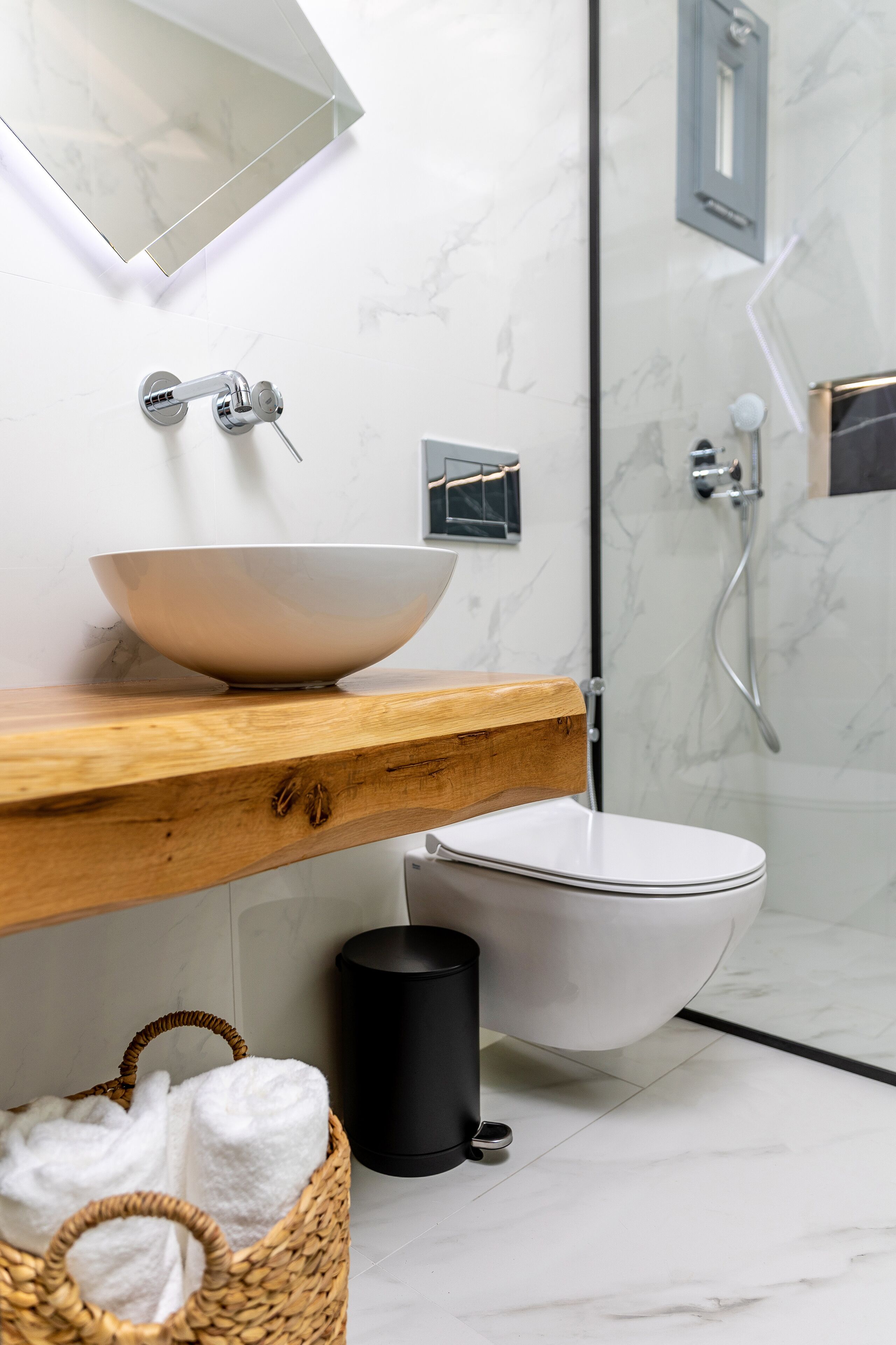 A modern bathroom with a wooden vanity, a white sink, a white toilet, a black trash bin, and a wicker basket with towels.