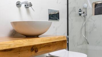 A modern bathroom with a wooden vanity, a white sink, a white toilet, a black trash bin, and a wicker basket with towels.