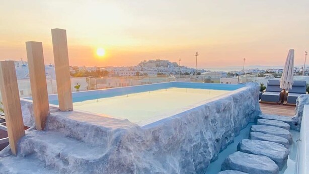A rooftop pool with stone steps and lounge chairs, overlooking a cityscape at sunset. - Alenor City Hotel (Naxos)