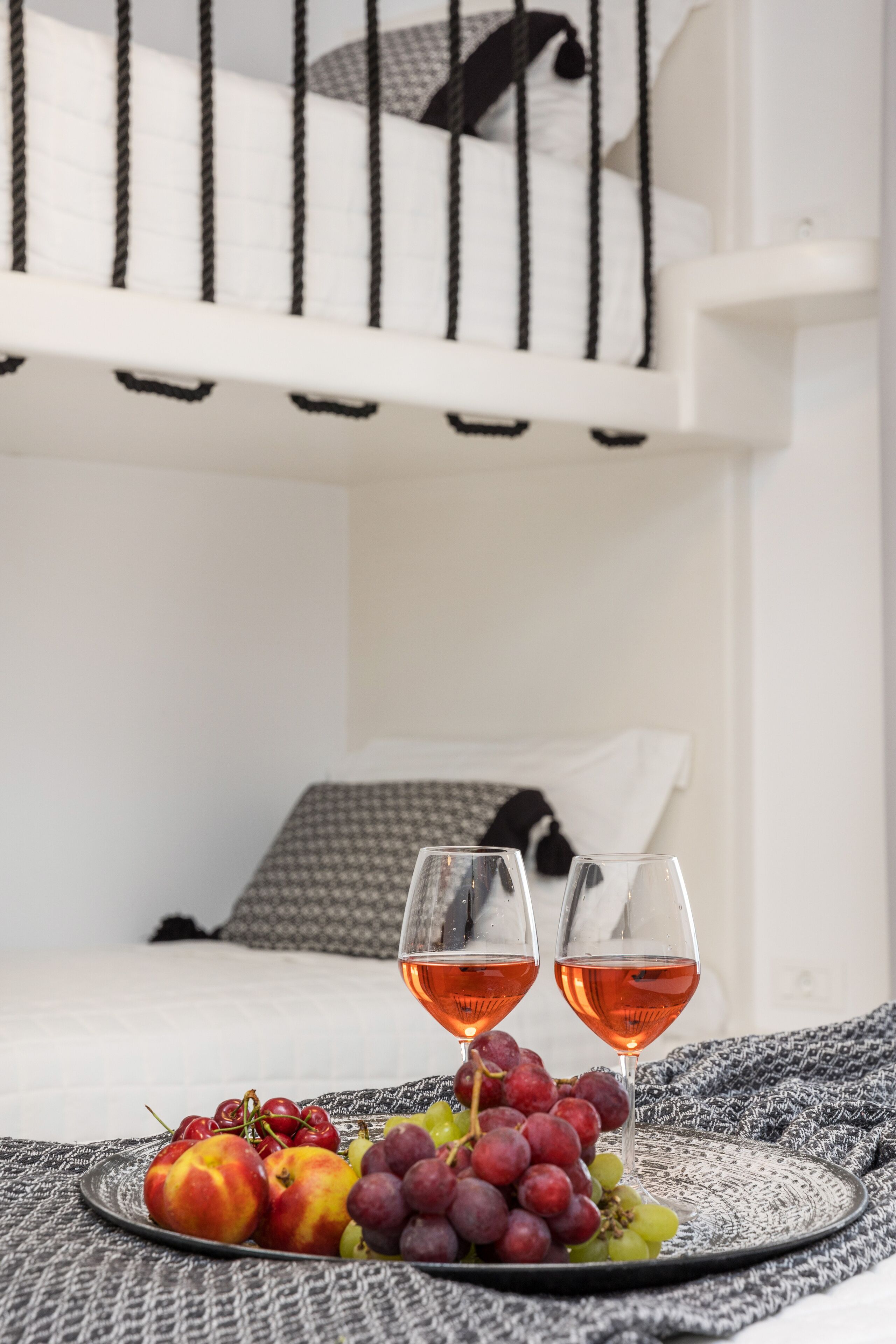 A bed with a white headboard and black vertical lines, a plate with grapes and peaches, and two glasses of red wine on a textured cloth.