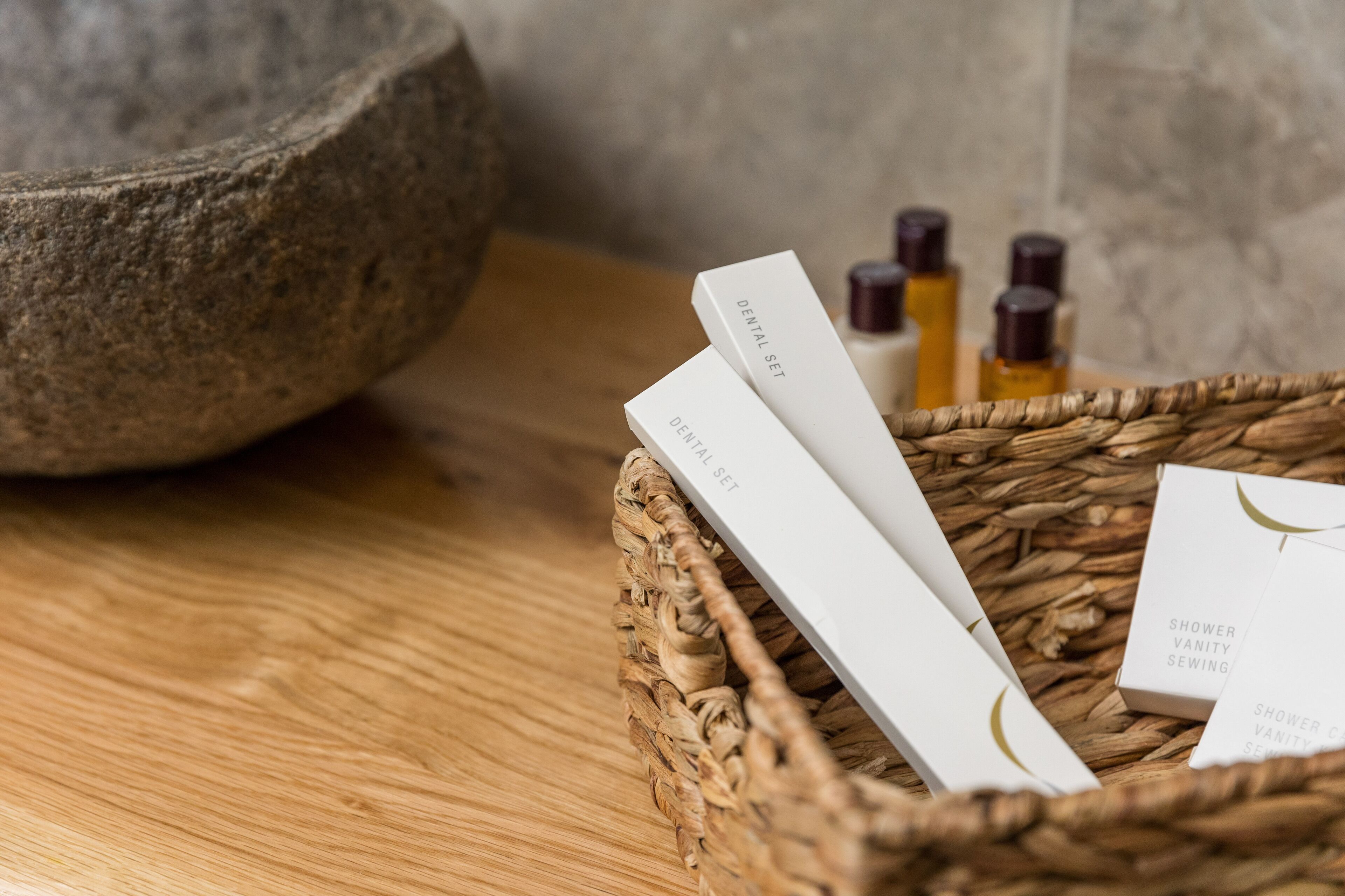 Un panier en osier contenant un nécessaire de soins dentaires et un ensemble d’accessoires pour la salle de bain, avec des bouteilles en arrière-plan.