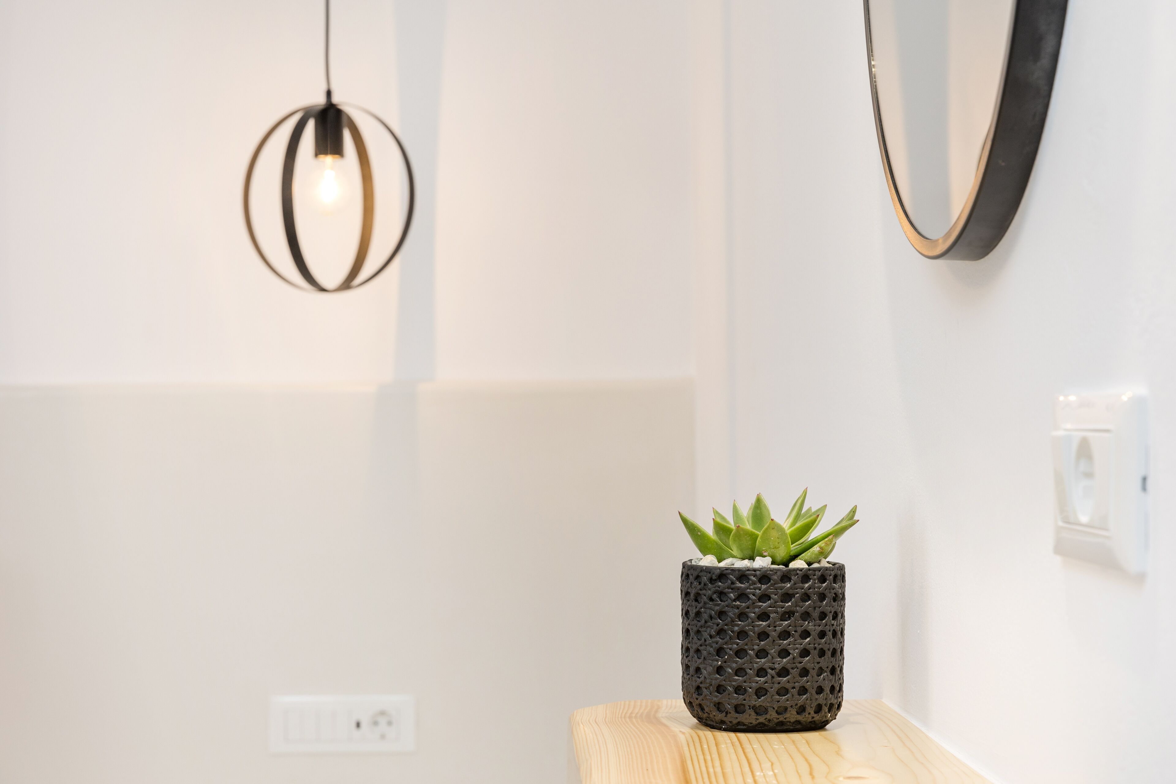 A modern interior with a wooden table, a textured black pot with a succulent, a pendant light, and a wall-mounted mirror.