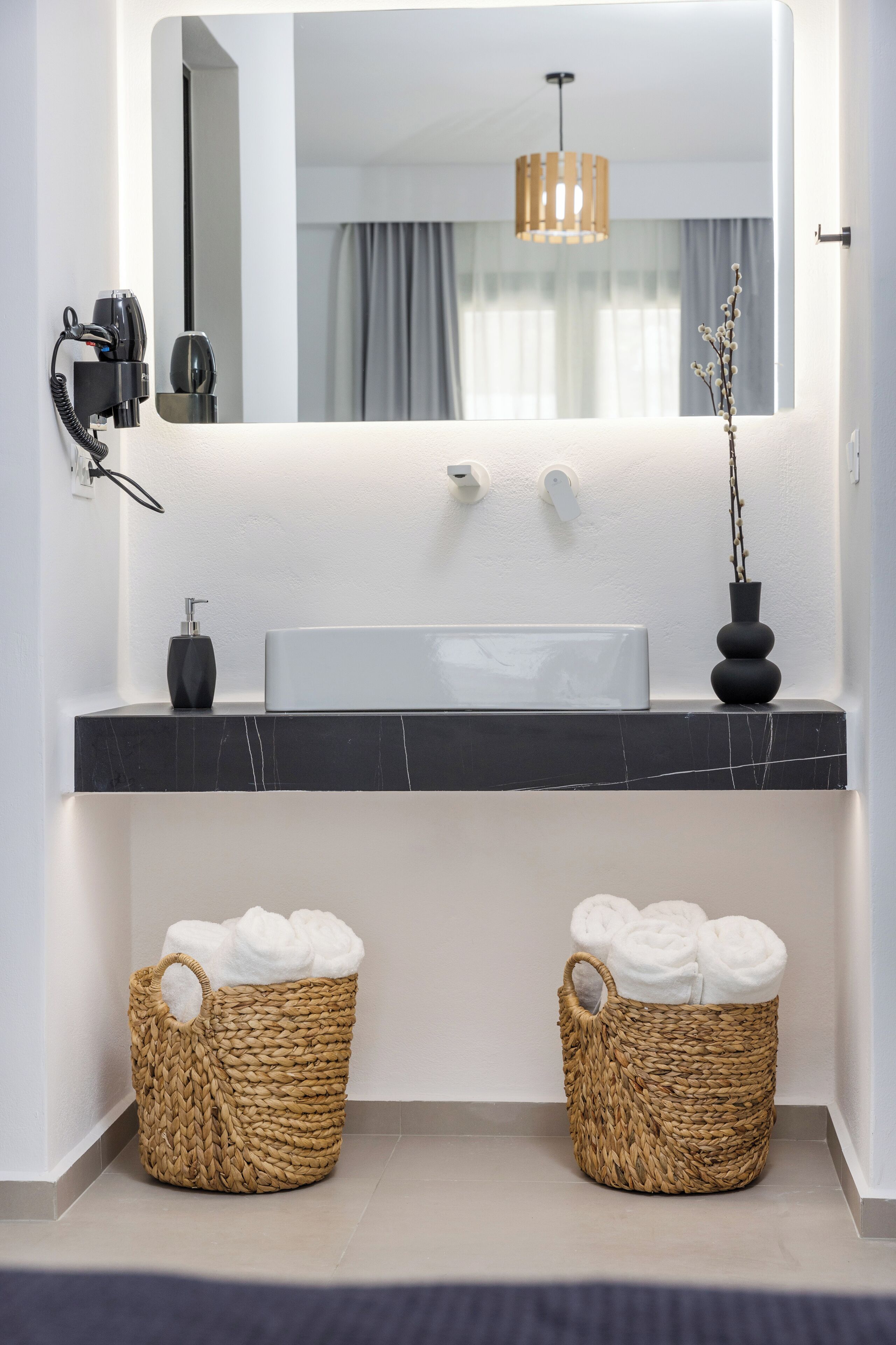A modern bathroom with a large mirror, a black marble countertop, a white sink, and two wicker baskets with towels.