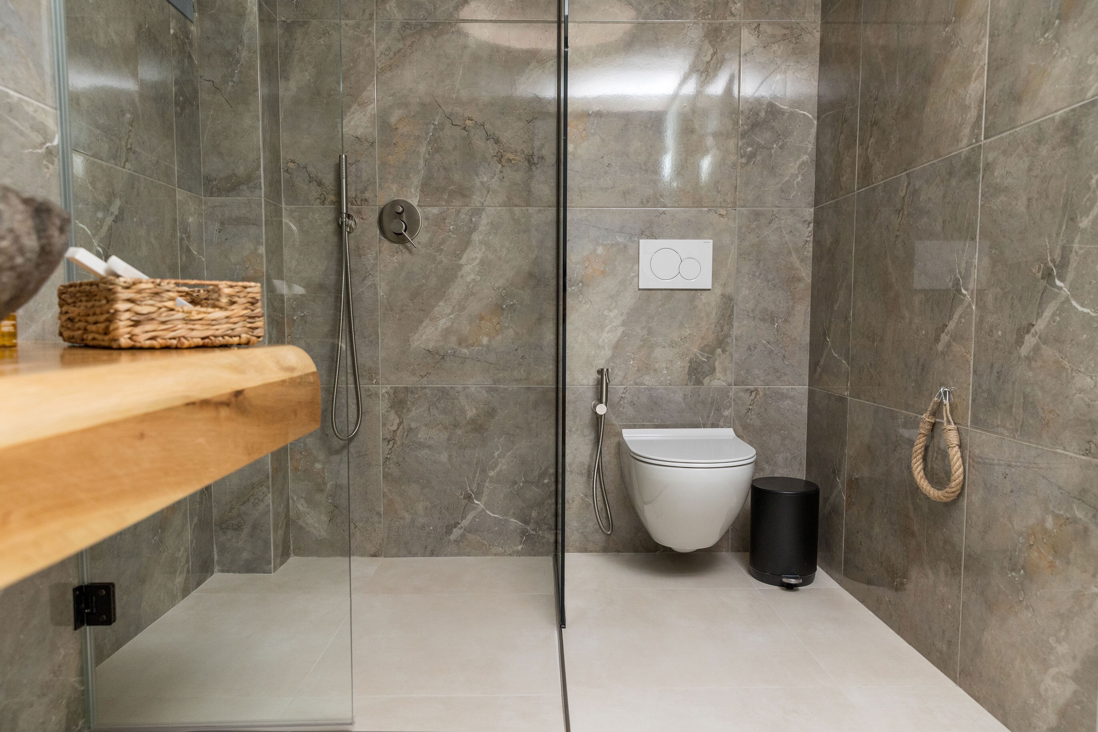 Ein modernes Badezimmer mit Duschkabine aus Glas, einem Holzregal, einer weißen Toilette und einem schwarzen Mülleimer.