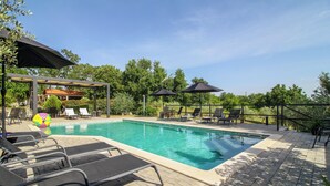 Ein rechteckiger Swimmingpool mit Liegestühlen und Sonnenschirmen, umgeben von viel Grün und blauem Himmel.