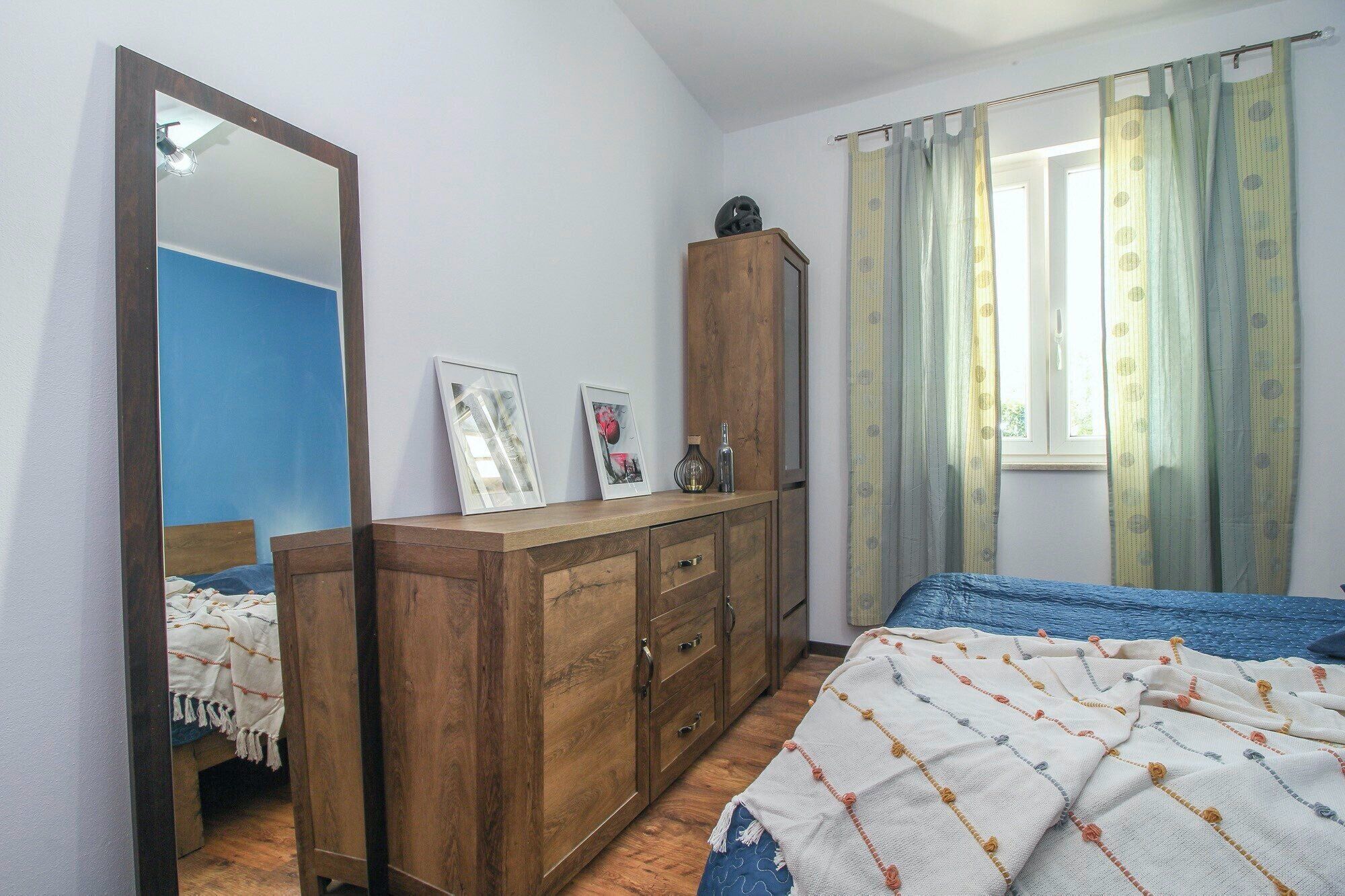 Une chambre à coucher avec une armoire en bois, un lit recouvert d’une couverture à motifs, un miroir et une fenêtre avec des rideaux.