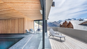 Un espace piscine extérieur moderne, doté d’une terrasse en bois, de chaises longues et offrant une vue sur des montagnes enneigées.