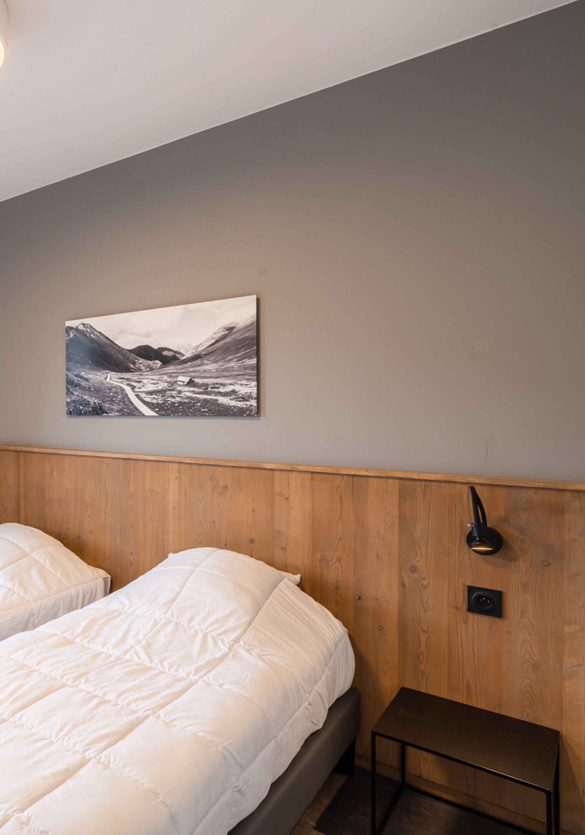 A bedroom with two beds, a wooden headboard, a bedside table, and a wall-mounted lamp.