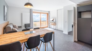 Un espace de vie moderne comprenant une table à manger en bois, des chaises noires, un canapé et une grande fenêtre offrant une vue sur les montagnes.