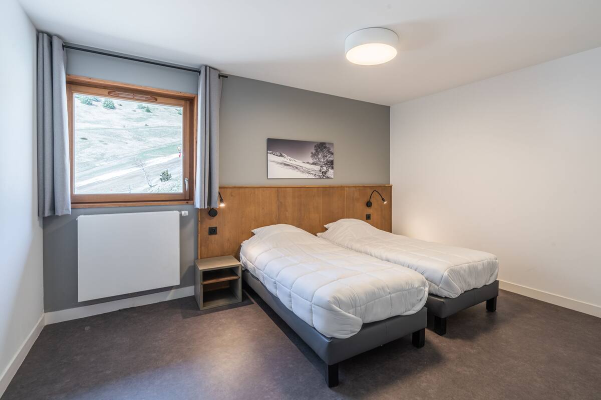 Une chambre avec deux lits simples, une fenêtre donnant sur un paysage enneigé, un radiateur et une petite étagère en bois.