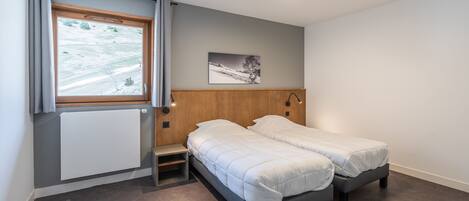 Un dormitorio con dos camas individuales, una ventana con vistas a un paisaje nevado, un radiador y un pequeño estante de madera.
