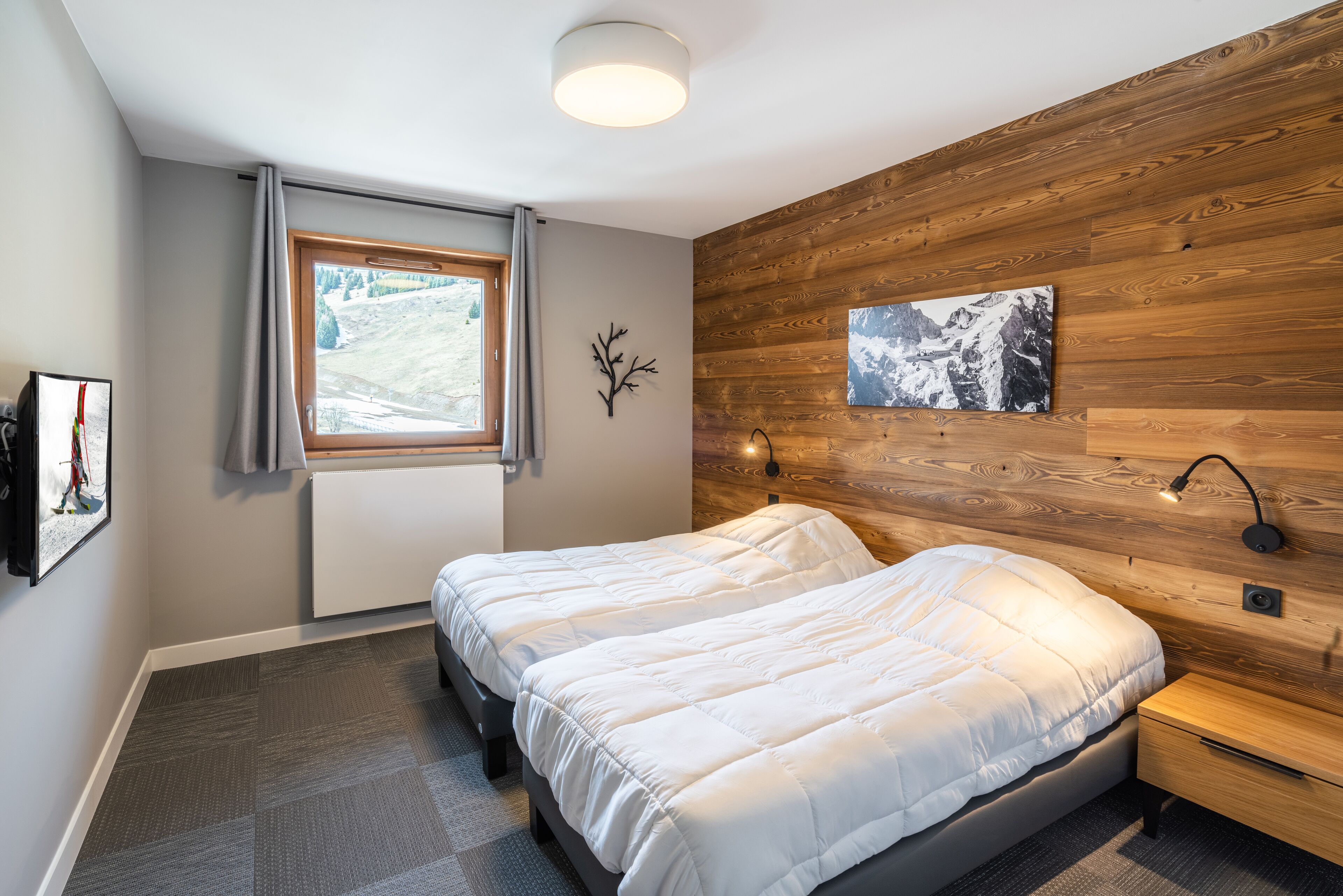Une chambre avec deux lits, une fenêtre donnant sur un paysage enneigé, un lambris en bois sur le mur et un tableau accroché.