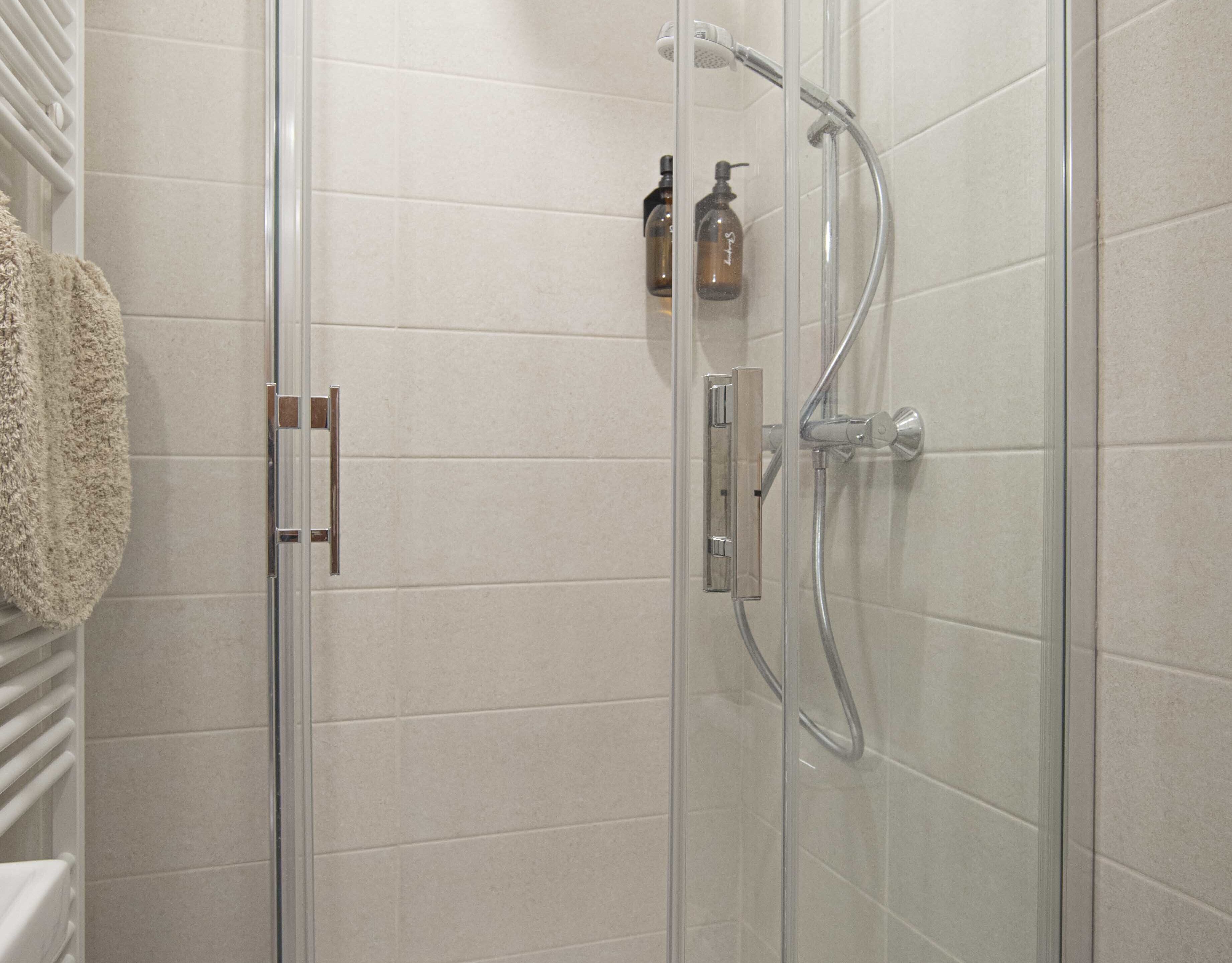 Een douche met glazen wand, een handdouche en een muurbekleding. Twee flessen shampoo en conditioner staan op de wand.