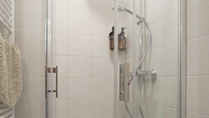 Une douche en verre avec un pommeau de douche à main et un robinet mural. Deux bouteilles de shampoing et d'après-shampoing sont fixées au mur.