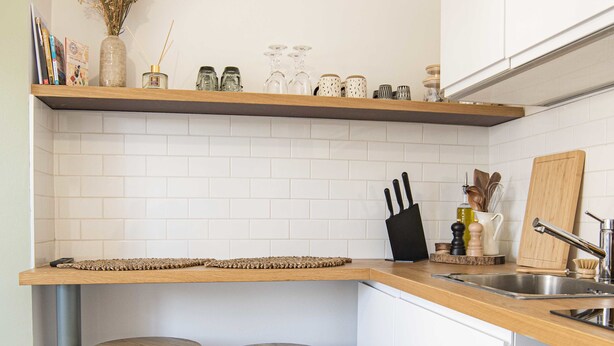 Une cuisine avec des carreaux blancs, des plans de travail en bois, deux tabourets, un bloc à couteaux et un évier.